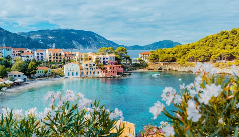 Vista panoramica del villaggio di Assos a Cefalonia, Grecia. Fiori bianchi e luminosi in primo piano sulla baia calma e turchese del Mar Mediterraneo e belle case colorate sullo sfondo.